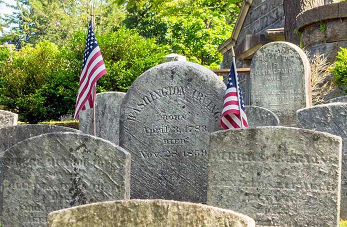 Several historic gravestones, including that of Washington Irving, with two small American flags in the ground at Sleepy Hollow Cemetery.