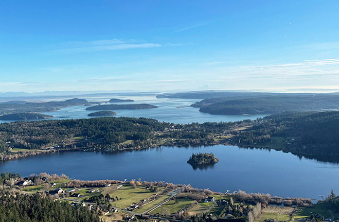 An aerial view of Anacortes and its stunning water scenery. The town contains several buildings and thick forests under a bright blue sky.