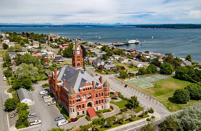 An aerial view of Port Townsend, focusing on the County Courthouse, a large, orange brick building with a clocktower on top and gold spires on each corner of the roof. There are several buildings and trees behind the courthouse, and the waters of Puget Sound and the Strait of Juan de Fuca are in the distance. 