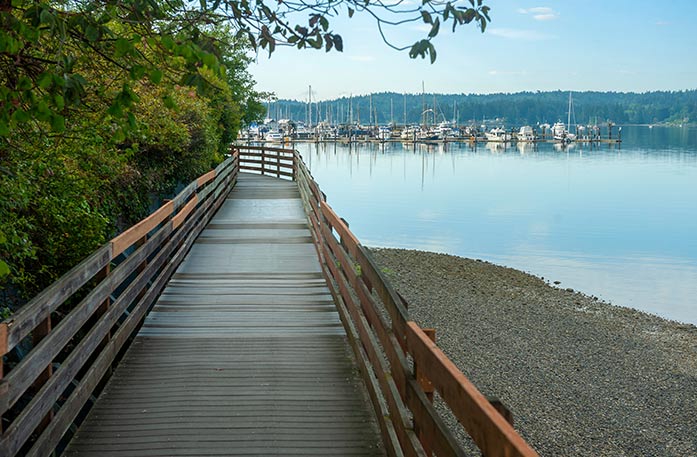 A wooden boardwalk in Liberty Bay Waterfront Park, alongside the bright blue waters of Puget Sound in Poulsbo. There are several sailboats docked on the water in the distance, and the left side of the boardwalk is lined with trees.