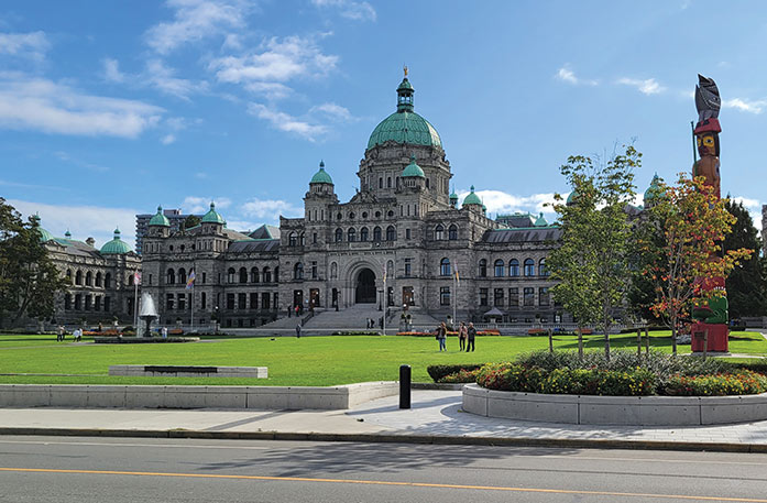 A road-side view of the Parliament Buildings, a large combination of castle-like buildings with a large, green spire on the center of the roof and smaller spires along the roof and several stairs leading to the archway in Victoria, BC. There is a grass lawn in front of the building with a water fountain in the center. By the edge of the road, there is a circular garden with flowers and small trees