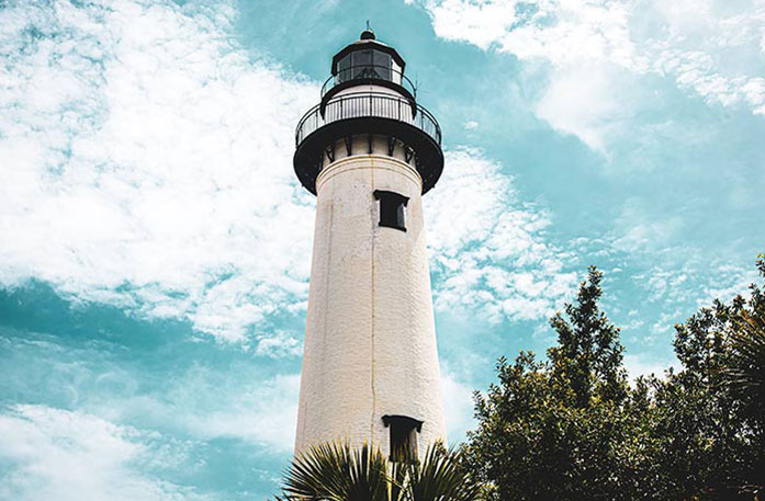 A bottom-up view of Amelia Island Lighthouse, a white lighthouse with a black balcony at the top under a cloudy blue sky.