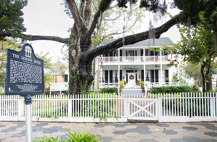 The Lesesne House, a small white house with a white picket fence and closed gate in front, separating the grass yard from the stone sidewalk in Amelia Island. There is a large live oak tree in the yard and a black sign on a metal pole that reads "The Lesesne House" with a lot of small writing below. 