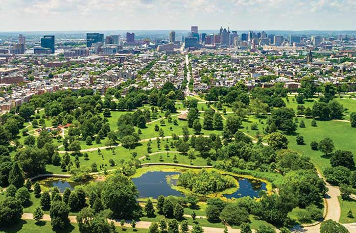 An aerial view of Baltimore, including a large pond surrounded by a forest of trees in the foreground, and countless buildings and skyscrapers in the background.