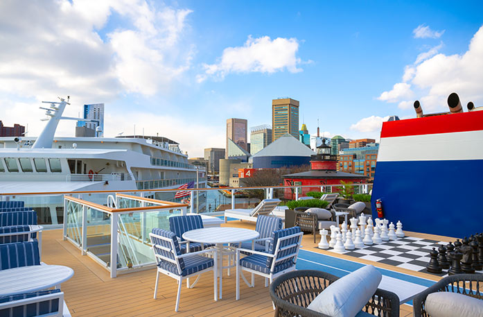 A view from the sun deck of an American Cruise Lines Coastal Cat, with American Constitution beside it and the city of Baltimore in the distance. There are a few chairs and tables on the sun deck, a staircase leading to the deck below, and a life-size chess board on the floor.