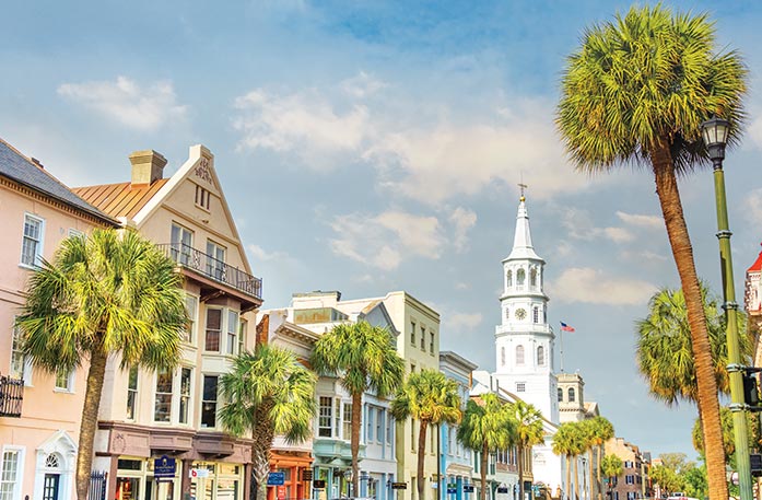 An endless line of colorful buildings and tall palm trees along a street in Downtown Charleston, including the tall, white spire on top of St. Michael's church.  