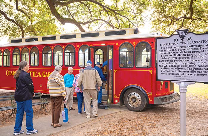 A group of people boarding a red trolley with yellow details, including "Charleston Tea Garden" written on the side, traveling to the Charleston Tea Garden. There is a sign to the right of the trolley with a long description of the Charleston Tea Garden and trees in the distance.