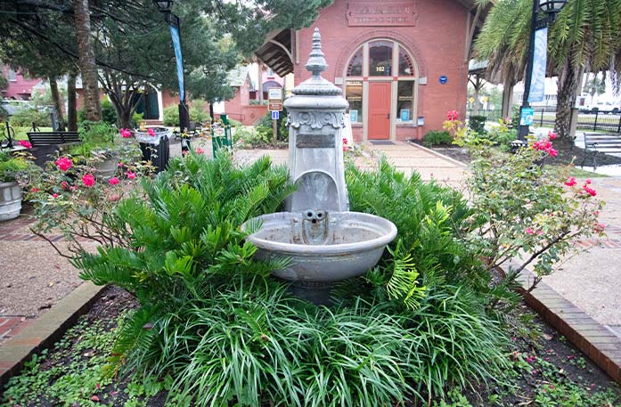 A small, cement colored fountain surrounded by plants in a small garden in Downtown Fernandina Beach. There is a small red building, park benches, and palm trees in the background.