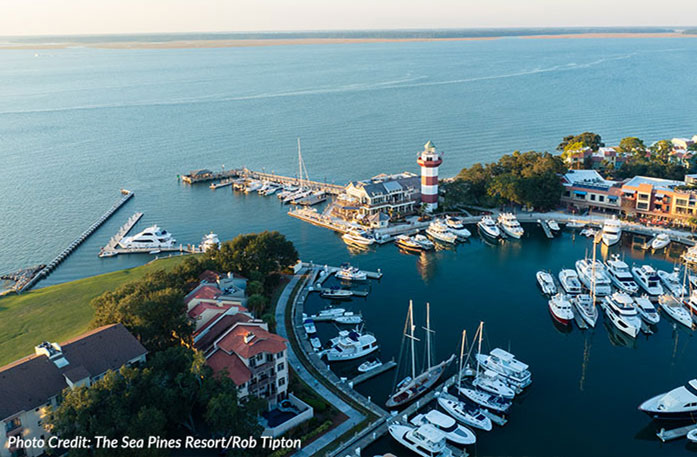 An aerial view of the Sea Pines Resort on Hilton Head Island with a circular marina with several small, docked boats. Along the dock, there is a red and white striped lighthouse. The marina leads to the Atlantic Ocean and a sand bar in the distance. There is a disclaimer in the bottom lefthand corner reading "Photo Credit: The Sea Pines Resort/Rob Tipton".