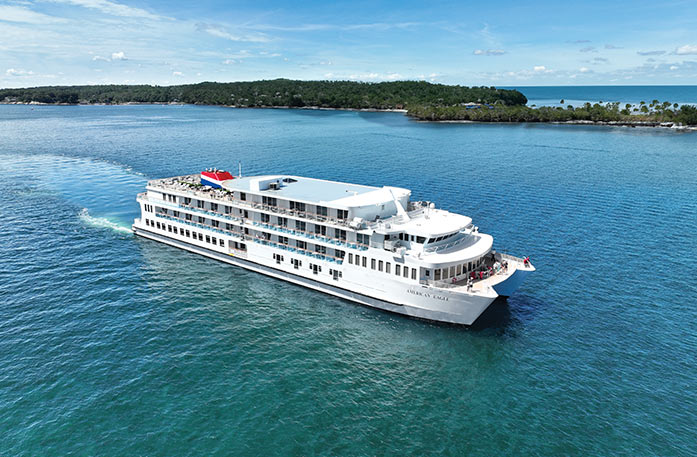 American Eagle Coastal Cat, a small white cruise ship sailing along the bright blue Gulf Coast waters with thick forest land in the distance.