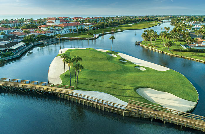 An aerial view of the Jacksonville cityscape, showcasing a large putting green on Ponte Vedra Inn's island green in the center of Lake Vedra. On the left side of the river, there is a large white building with an orange roof, Ponte Vedra Inn. There are palm trees surrounding the green and the shoreline.