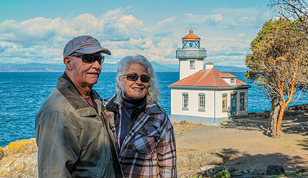 A couple stands together smiling for a photograph taken of them in front of the bright blue Puget Sound with a small lighthouse on the shore.