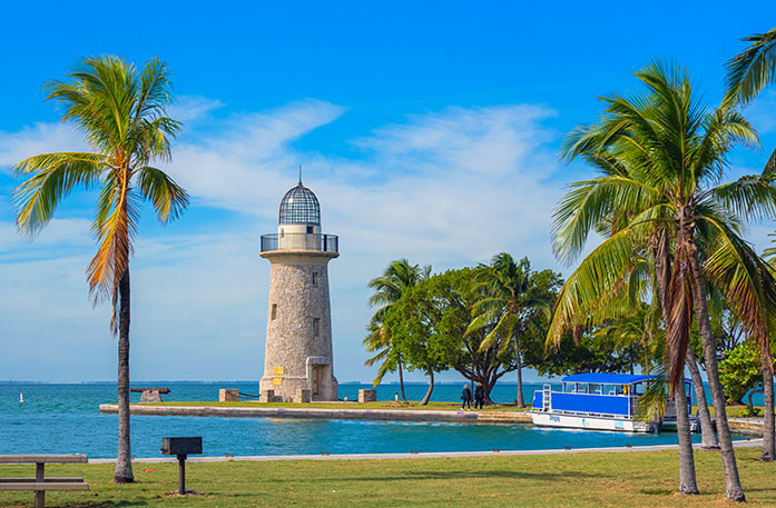 Boca Chita Key, a stone lighthouse with a wraparound black balcony at the top, almost completely surrounded by water besides a narrow path to land in Key Biscayne. There are a few palm trees in the foreground, and there is a vibrant blue boat docked on the narrow path.