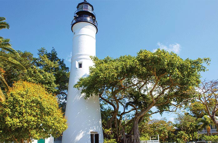 A bottom-up view of Key West Lighthouse & Keeper's Quarters, a white lighthouse with oak trees standing beside it under a blue sky at the Key West Art & History Museum.