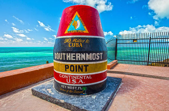 A buoy structure at the southernmost part of Continental U.S.A. beside the water in Key West, Florida. It is separated into different colors by white lines, starting with red, then black, yellow, red, and black. Each section has words on it in different colors, ultimately reading "The Conch Republic, 90 Miles to Cuba. Southernmost Point Continental U.S.A. Key West, FL. Home of the sunset."