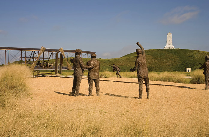 A recreation of the first flight, with a large sculpture of an airplane with a statue of a person laying on it, and four statues on the ground behind it at Wright Brothers National Memorial in Kitty Hawk.