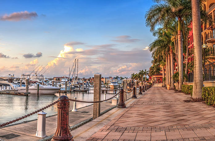 A brick path alongside the Gulf of Mexico, where several boats are docked. On the right side of the path, there are tall, red and orange buildings with palm trees in front of them. 