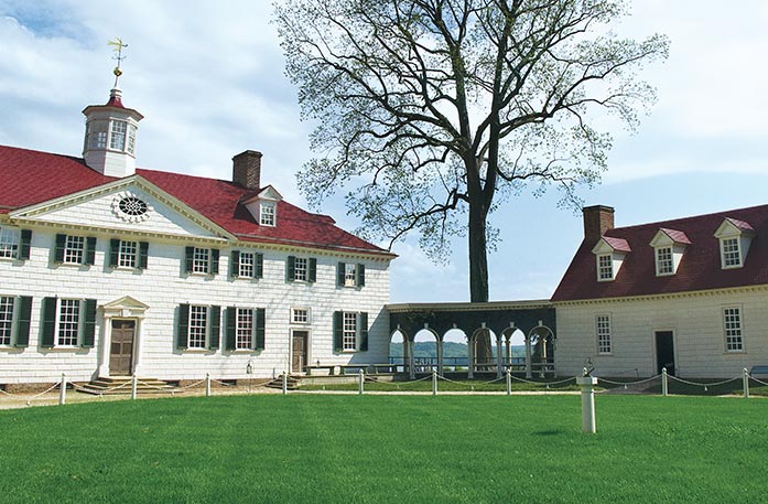 George Washington's Estate in Mount Vernon, a large white house with a red roof and green shutters is to the left of a smaller white house with a red roof, with an breezeway connecting the two.