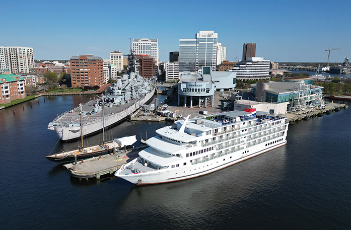 An American Cruise Lines small cruise ship docked in Norfolk, with an aerial view of the cityscape. To the left of the cruise ship, the large Battleship Wisconsin is docked.