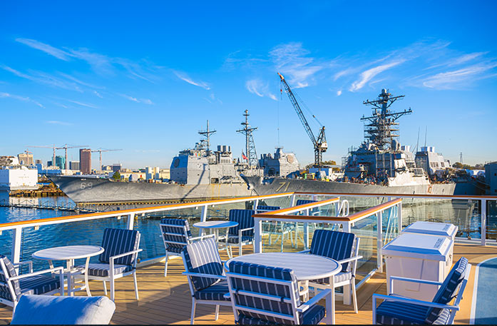 A view from the sun deck of an American Cruise Lines Coastal Cat of two large aircraft carrier ships, USS Dwight D. Eisenhower and USS Theodore Roosevelt, docked in the Norfolk Naval Station. On the sun deck, there are several blue and white striped chairs and white round tables, surrounded by a glass railing.