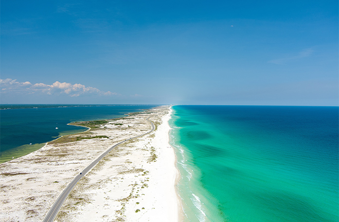 A roadway between the sand of Pensacola Beach, dividing the clear blue waters of Pensacola Bay.