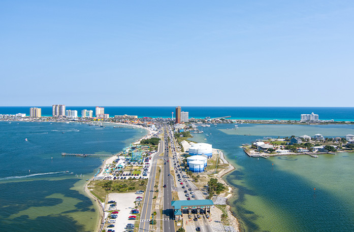 An aerial view of Pensacola, where a highway surrounded by buildings and greenery divides the clear blue waters of Pensacola Bay. In the distance, the highway splits in two with the Gulf Coast in the distance. 