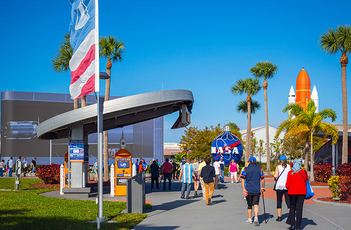 NASA's Kennedy Space Center in Port Canaveral with several visitors walking on the gravel paths towards the building. There is a blue life-size ornament  with the "NASA" logo in large white letters, and an orange and white spaceship partially covered by a palm tree in the distance.