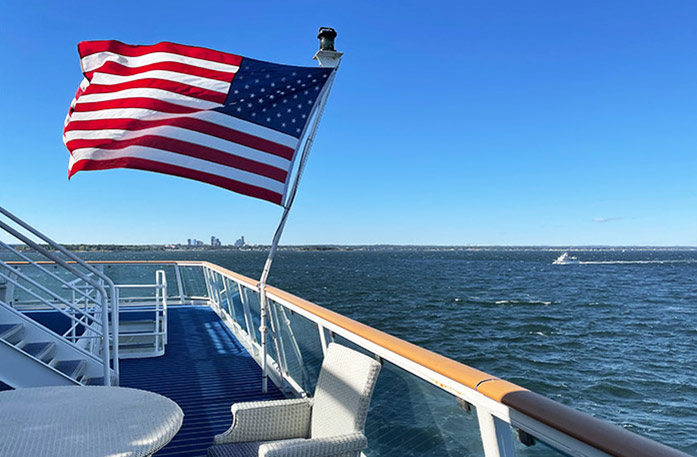 On the deck of an American Cruise Lines Coastal Cat, there is a large American flag, with its pole on the glass railing, blowing in the wind with the views of the Intracoastal Waterway. The floor of the deck is dark blue, a few stairs can be seen to the left, and there is an empty tan colored chair and table in the foreground.