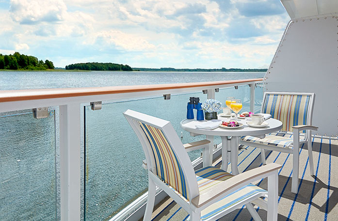 On an American Cruise Lines Coastal Cat private balcony, two blue and yellow striped chairs surround a round, white table with a two plates of food and yellow drinks, binoculars, and a pot with blue flowers. Outside the glass railing is the Intracoastal Waterway.