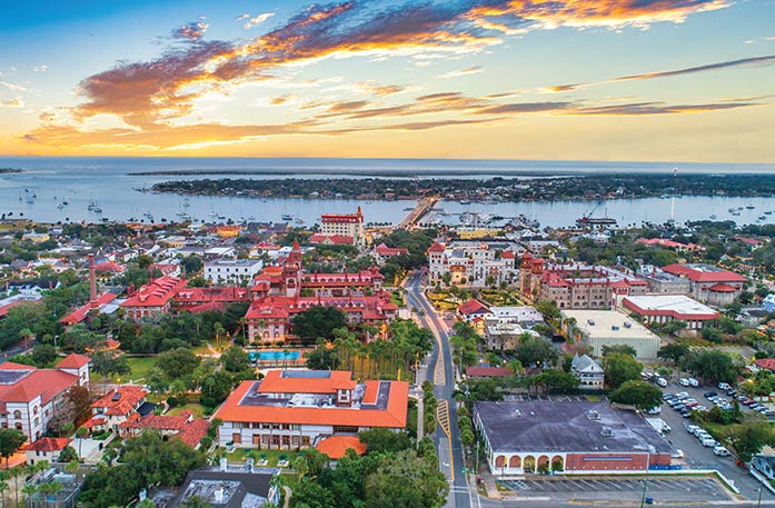 An aerial view of the St. Augustine cityscape under an orange sunset over the distant bay. Most of the buildings have orange or red rooftops, creating a vibrant scene.