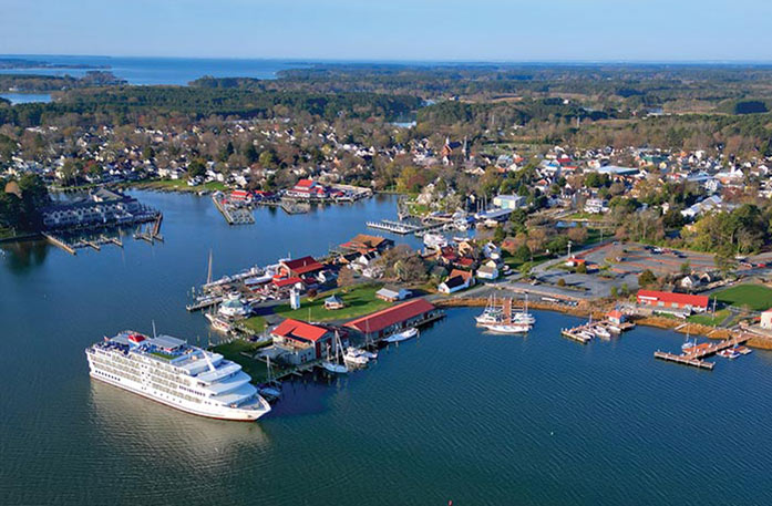 An aerial view of an American Cruise Lines small cruise ship docked in St. Michaels along the Chesapeake Bay with trees and buildings filling the shore.