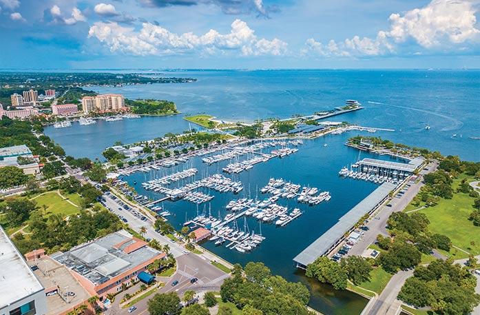 An aerial view of boats docked in a marina in St. Petersburg with the bright blue Gulf Coast waters spanning for miles in the distance. There are several buildings and trees on the shoreline.