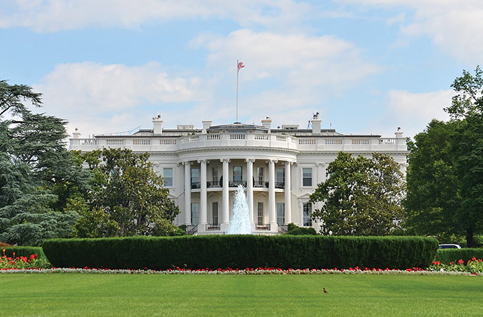 The White House, a white historic building with tall pillars that form a half circle under a balcony. There is a tall flag pole on the roof with an American flag on top. In front of the house, there is a water fountain with trimmed bushes, trees, and small red flowers surrounding it. 