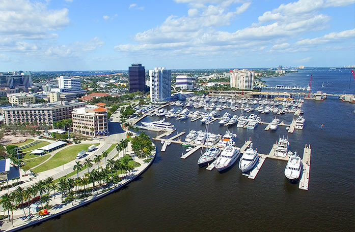 Aerial view of boats docked in the deep blue Florida coast marina alongside the West Palm Beach cityscape with several tall buildings and palm trees.