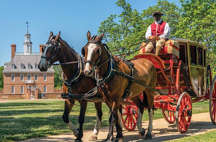 Two brown horses are pulling a red carriage with a person dressed in colonial clothing sitting on top of the carriage in Williamsburg. Governor's Palace, a two-story brick building with tall, narrow windows on both stories and the grey roof, two chimneys on each side, and a spire in the middle.