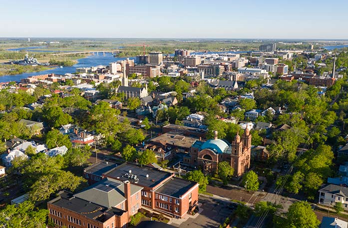 An aerial view of the city of Wilmington, highlighting its many buildings, trees, and the Cape Fear River in the distance.