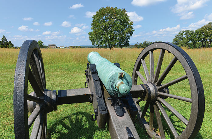 A green cannon with two large wheels on the grounds of the American Revolution Museum at Yorktown. The cannon is facing a large field with tall grass and one large tree in the middle.