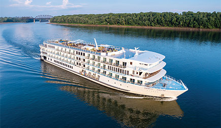 The American Riverboat American Serenade gliding along the Mississippi River, with the tree-filled shore and a bridge in the background.