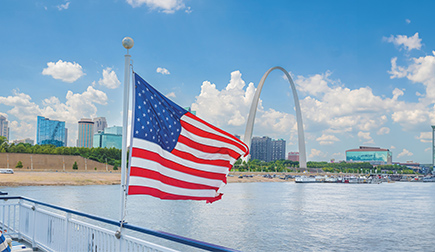 An American flag on a pole connected to the blue and white railing of an American Riverboat blows in the wind along the Mississippi River and the St. Louis Gateway Arch in the distance.