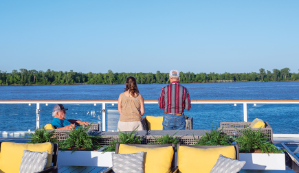 On the American Riverboat Sun Deck, a couple is standing against the glass railing gazing at the Mississippi River, the trees lining the horizon line, and the clear blue sky. There are four patio chairs with yellow and grey pillows in the foreground, and in between those and the couple, there is a man sitting on a chair facing the right.