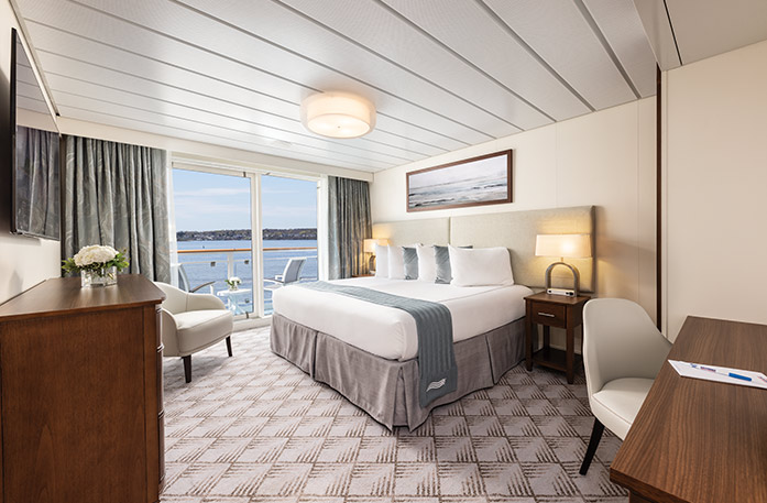 The Premium Balcony stateroom on American Legend, with floor-to-ceiling sliding glass doors that lead to a balcony with a table and chairs overlooking the Gulf Coast. There is a bed with white and grey pillows and a grey bed runner on the right side of the room, with a bedside table and a lamp on each side, and a painting of waves above it. The bed is facing a TV on the wall, above a dresser.