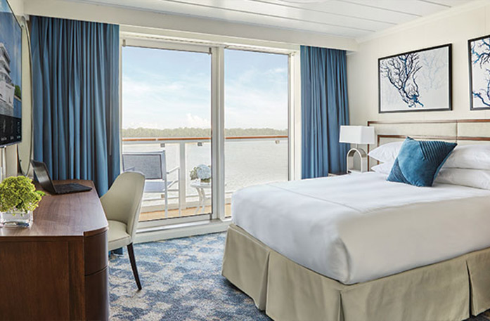 The Single Balcony stateroom on American Encore, with large floor-to-ceiling sliding glass doors, with blue curtains on each end, that lead to a private balcony with a table and chairs overlooking the Columbia River. There is a bed with white bedding and a blue accent pillow on the right side of the room, facing a wood desk and tan chair and a TV on the opposite wall.