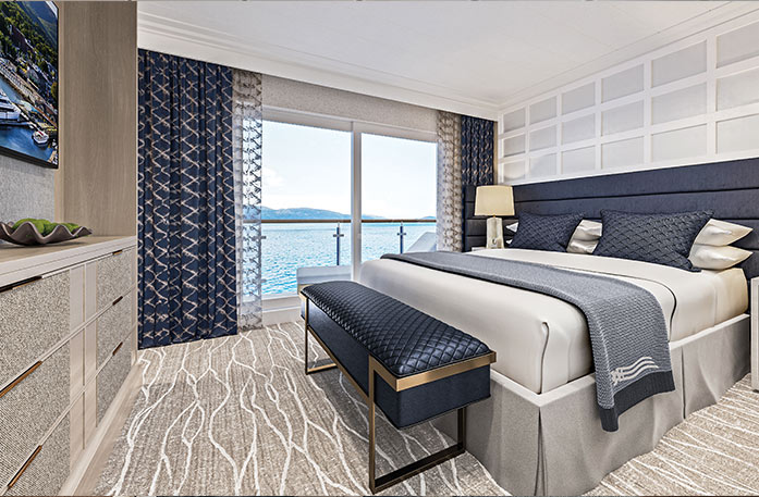 The Sky Suite on American Encore, with large floor-to-ceiling sliding glass doors leading to a private balcony overlooking the Columbia River. There is a bed against the wall on the right side with navy blue pillows, tan bedding, a grey bed runner, and a navy blue and gold bench at the foot of the bed. There is a dresser against the wall across from the bed and a TV on the wall above it.