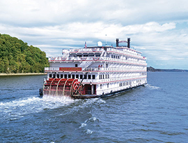 American Heritage, an American Cruise Lines Paddlewheeler decorated with patriotic red white and blue bunting flags on the back railings, sailing along the Mississippi River. The back of the small cruise ship has a large, red paddlewheel, and the shore on the left side of the river is covered in trees.