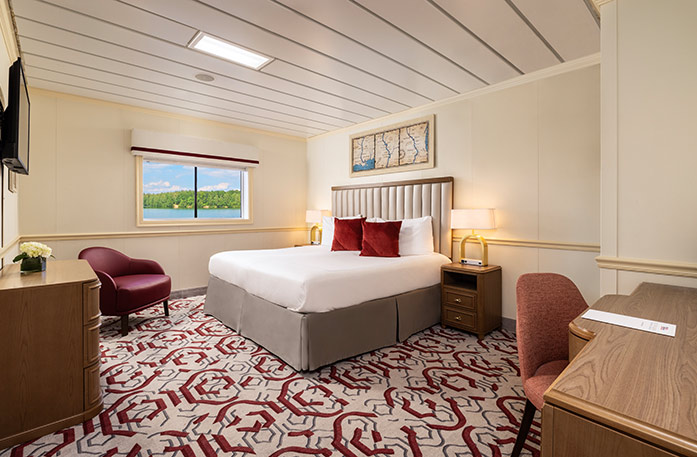 The Standard Waterview stateroom on American Heritage, with a large picture window overlooking the Mississippi River. There is a bed on the right side of the room with two red accent pillows, and end tables with a lamp on each side of the bed. There is a wooden dresser and a TV on the wall above it across from the bed, and a red chair in the corner.