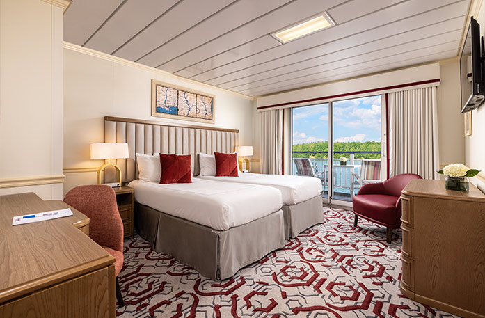 The Deluxe Balcony 1 stateroom on American Heritage, with floor-to-ceiling sliding glass doors that lead to a private balcony with a table and chairs overlooking the Mississippi River. There are two beds beside each other on the left side of the room with a red accent pillow on each, across from a wooden dresser and a TV on the wall above it across from the bed, and a red chair in the corner.
