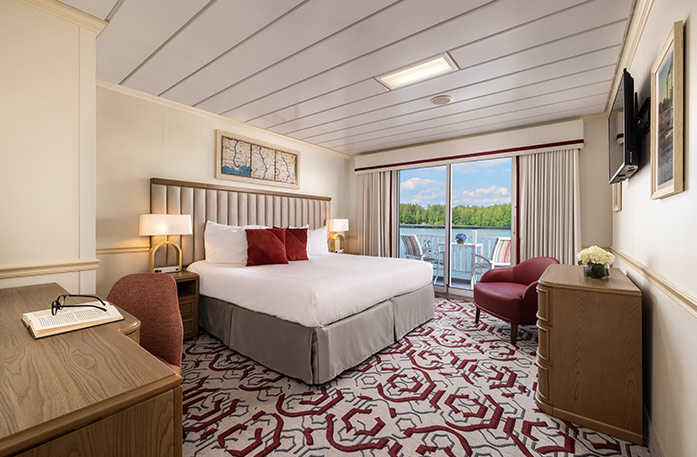 The Deluxe Balcony 2 stateroom on American Heritage, with floor-to-ceiling sliding glass doors that lead to a private balcony with a table and chairs overlooking the Mississippi River. There is a bed on the left side of the room and end tables with a lamp on each side of the bed, across from a wooden dresser and a TV on the wall above it across from the bed, and a red chair in the corner.