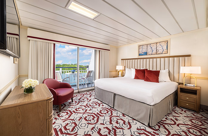 The Premium Balcony stateroom on American Heritage, with floor-to-ceiling sliding glass doors that lead to a private balcony with a table and chairs overlooking the Mississippi River. There is a bed on the right side of the room and end tables with a lamp on each side of the bed. There is a wooden dresser and a TV on the wall above it across from the bed, and a red chair in the corner.
