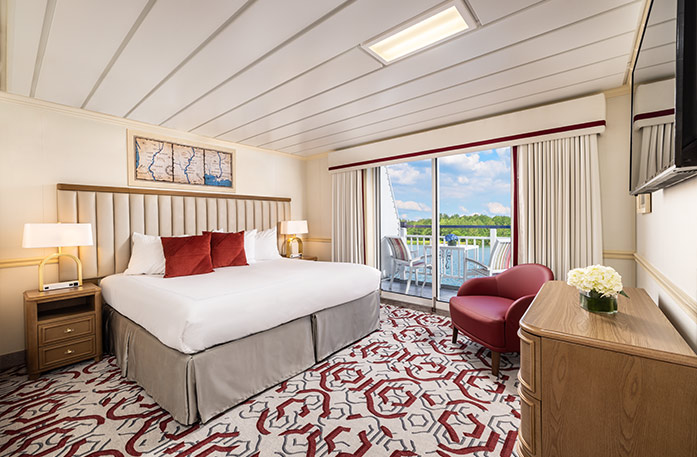 The Standard Balcony stateroom on American Heritage, with floor-to-ceiling sliding glass doors that lead to a private balcony with a table and chairs overlooking the Mississippi River. There is a bed on the left side of the room and end tables with a lamp on each side of the bed, across from a wooden dresser and a TV on the wall above it across from the bed, and a red chair in the corner.