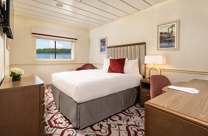 The Single Waterview stateroom on American Heritage, with a large picture window overlooking the Mississippi River. There is a bed on the right side of the room with a red accent pillow, and an end table with a lamp on one side of the bed. There is a wooden dresser and a TV on the wall above it across from the bed, and a red chair in the corner.